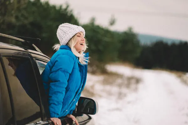 Žena v zimnom oblečení vychýlená z auta počas cesty na zimnú dovolenku v zasneženej krajine.